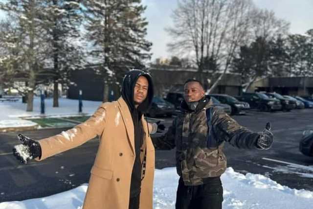 Deux jeunes hommes souriants, bras ouverts, dans la neige devant des sapins.