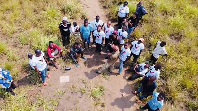 Vue aérienne d’une trentaine de personnes, beaucoup en t-shirt blanc Variable Fly, avec des habitants, sur un chemin de terre au milieu des herbes.